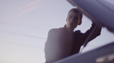 Genç, sıska bir Avrupalı adam arabasını tamir eder ve kaputu kapatır.