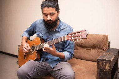 Gündelik giysiler içinde gitar çalan, bir odada koltukta oturan bir adam.