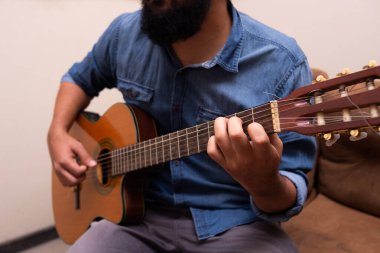 Gündelik giysiler içinde gitar çalan, bir odada koltukta oturan bir adam.