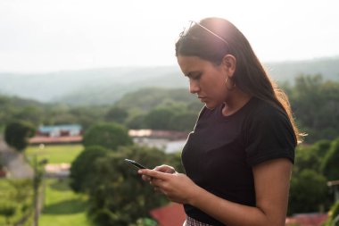 Cep telefonuna yazılar yazan genç bir kız, arka planda dağlar ve evler var.