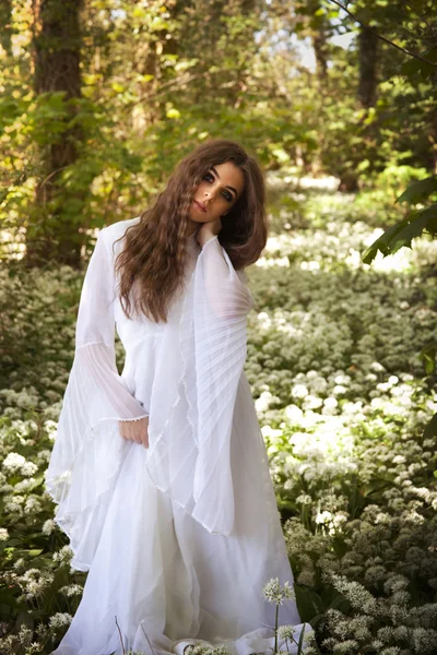 Hermosa mujer en vestido blanco largo de pie en un bosque en un capé de flores blancas — Foto de Stock