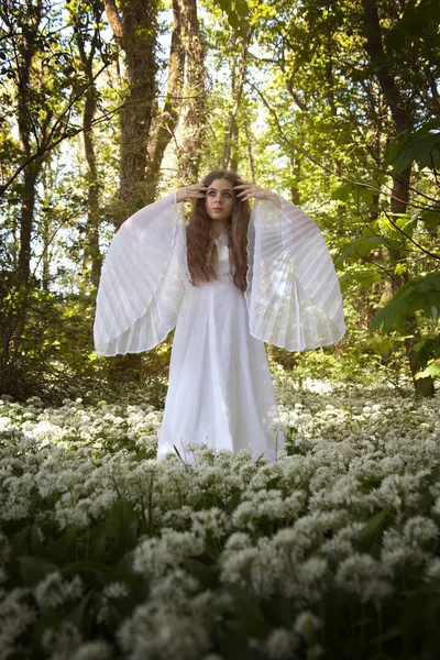 Hermosa mujer en vestido blanco largo de pie en un bosque sobre una alfombra de flores blancas — Foto de Stock