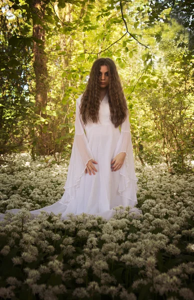 Hermosa mujer en vestido blanco largo de pie en un bosque sobre una alfombra de flores —  Fotos de Stock