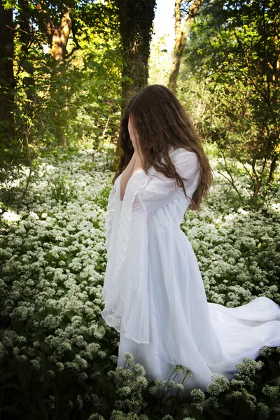 Mujer vestida con un largo vestido blanco de pie en un bosque cubriéndose la cara con las manos — Foto de Stock
