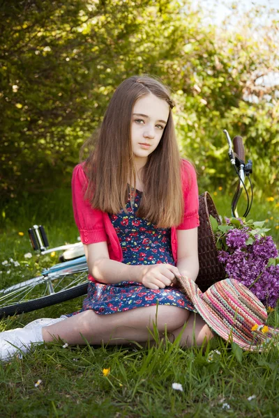 Vacker tonårsflicka som sitter i ett fält med en cykel med en liggande på gräset — Stockfoto