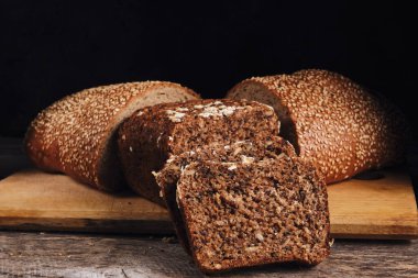 Cut in half a loaf sprinkled with sesame seeds and sliced cereal bread on a cutting board with a black background.