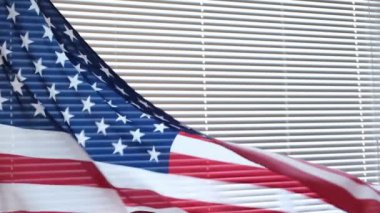 Perdelerle kaplı bir pencerenin arka planında rüzgarda sallanan Amerikan bayrağı.