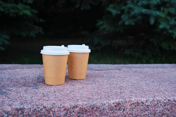 Two cups of coffee to take away in the park on a marble bench against a background of green spruce