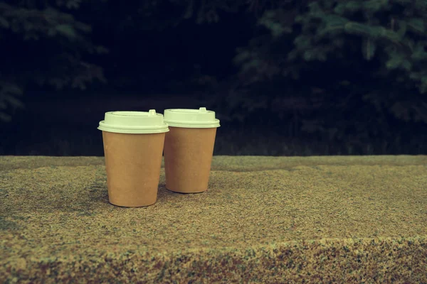 Two cups of coffee to take away in park on marble bench against background of green spruce at night