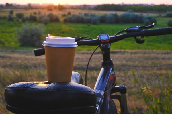 Glass coffee take away on bike seat against background green grass at sunset