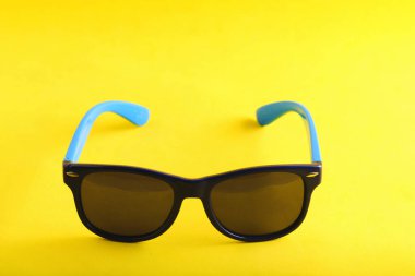 Mavi çerçeveli güneş gözlüğünü kolları desenli, sade sarı arka planda ortalanmış.