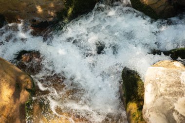 Kayalık bir derede yosun kaplı taşlar arasında akan beyaz suyun dinamik yakın çekimi. Köpüklü sprey hareket ve enerjiyi yakalar, doğa, seyahat ve açık hava yaşam temaları için mükemmeldir..