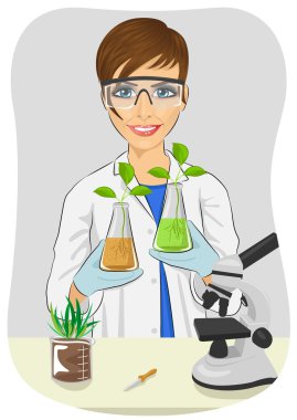 Young woman biologist in white coat holding two flasks with plants next to microscope in laboratory clipart