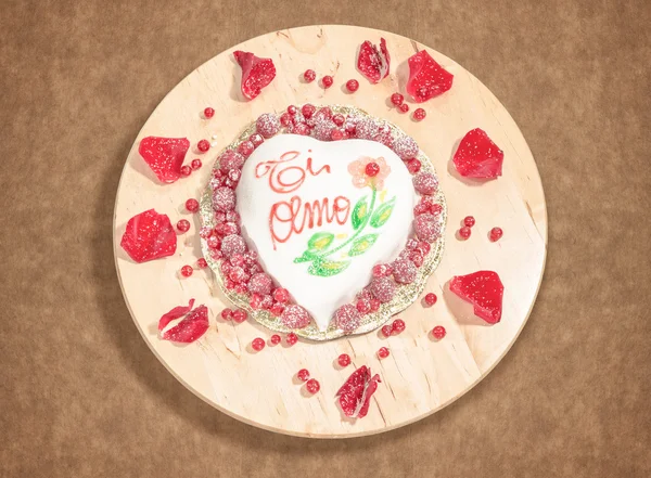 Valentine cake in the shape of heart decorated with raspberries,icing sugar and red rose petals,placed on a round wooden centerpiece. — Stock Photo, Image