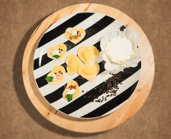 Black and white dish with handmade ravioli in the shape of heart,open and closed,fresh cheese and  few grains of black pepper,placed on a round centerpiece of wood. — Stock Photo, Image