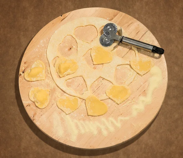 Raviolis caseros en forma de corazón, con masa cruda y cortadora de ruedas colocados en una pieza central redonda en madera . — Foto de Stock