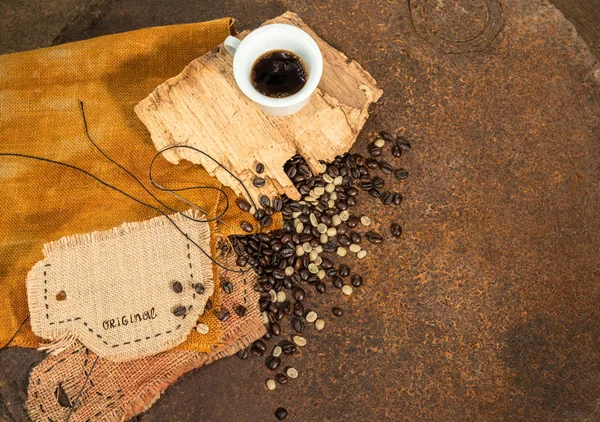 Eine Tasse Kaffee mit einer anderen in Jute genähten Tasse und ein paar Kaffeebohnen auf altem Holz und rostiger Textur. — Stockfoto