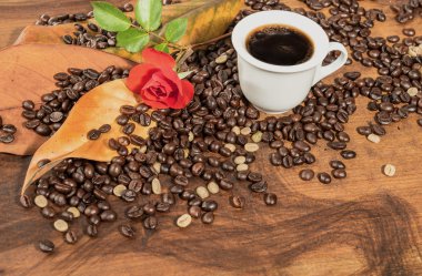 A cup of  black coffee with coffee beans,red rose and dry leaves of magnolia placed on wooden table.