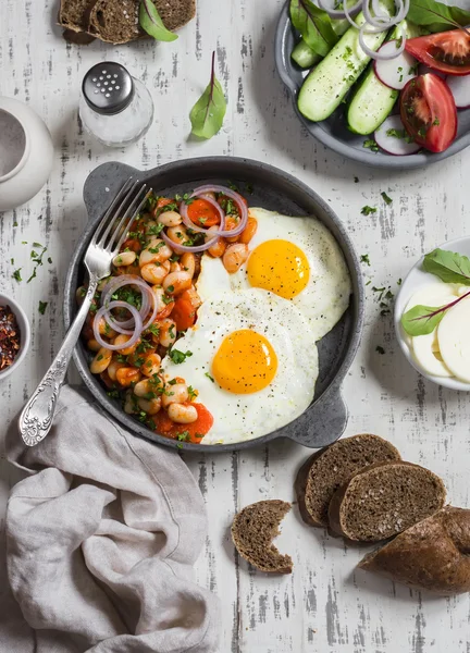 Delicious  breakfast or snack - a fried egg, beans in tomato sauce with onions and carrots, fresh cucumbers and tomatoes, homemade rye bread on light wooden background