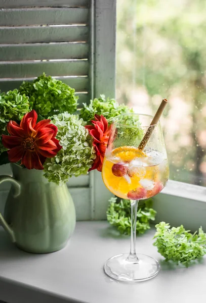 Orange alcohol ice cocktail and a bouquet of summer garden flowers in a vintage ceramic vase on the window on a summer day