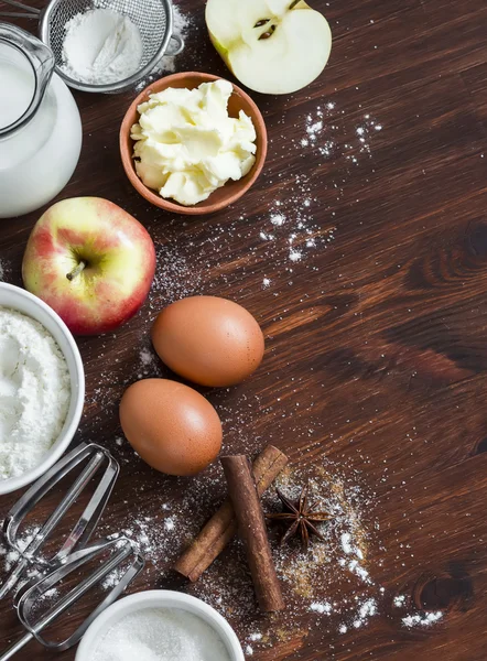 Ingredients and tools for baking - flour, eggs, butter, apples, cinnamon on a brown rustic wooden surface.