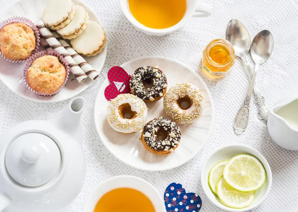 Valentine's day romantic Breakfast. Lemon green tea and sweets - banana muffins, cookies with caramel and nuts, donuts with chocolate and lemon glaze, tea set, red paper hearts on a white tablecloth on a light surface.