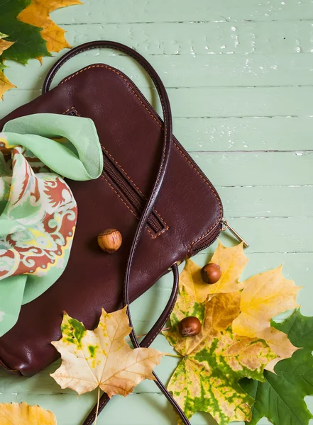 women\'s brown leather hand bag, silk neck scarf and yellow maple leaves on a light wooden surface