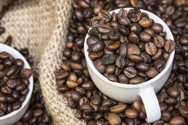 Xícaras cheias de grãos de café em um saco de serapilheira cheio de grãos de café — Fotografia de Stock