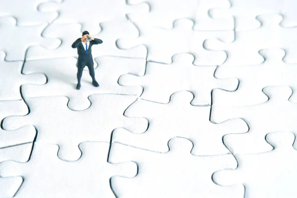 Businessmen standing above complete jigsaw puzzle using binoculars on wooden table. Miniature tiny people toys photography.