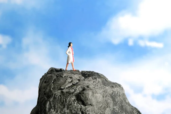 Miniature people toy figure photography. Superhero doctor concept. A women wearing white doctor coat or snelli standing above rock with blue cloudy sky background. Image photo