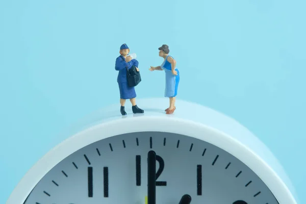 Miniature people toys conceptual photography. Delivery on time service. Postman courier standing above the clock, isolated on blue background.