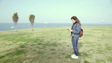 Genç öğrenci, bir kız güneşli bir günde deniz kenarındaki bir parkta bir şey arıyor, bir şey arıyor ve yoluna devam ediyor. Gerçek neşe ve duygu. Açık havada yürü. Yavaş çekim