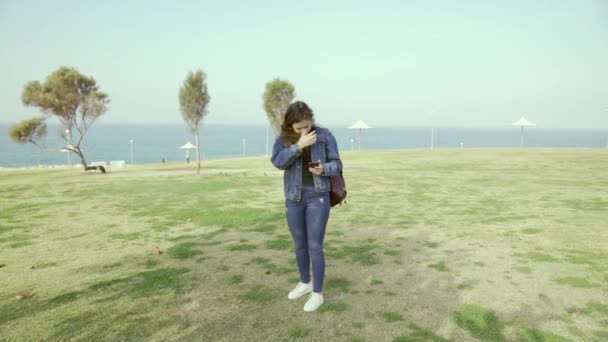 若い学生は 女の子が晴れた日に海のそばの公園にあり 電話でメッセージを入力します 電話で何か探してる オープンエアの中を歩く スローモーション — ストック動画
