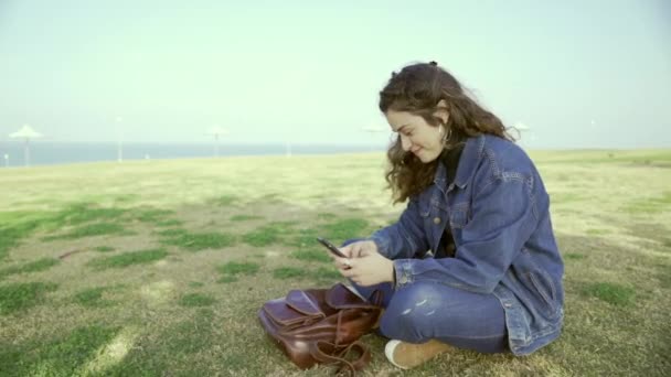 若い学生は 女の子が晴れた日に海のそばの公園にあり 地面に何かを探して座って メッセージを印刷します 本当の喜びと感情 オープンエアの中を歩く スローモーション — ストック動画