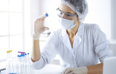 Koruyucu gözlüklü profesyonel kadın bilim adamı güneşli laboratuvarda reaktörlerle araştırma tüpü kullanıyor. Tıp ve bilim araştırmaları