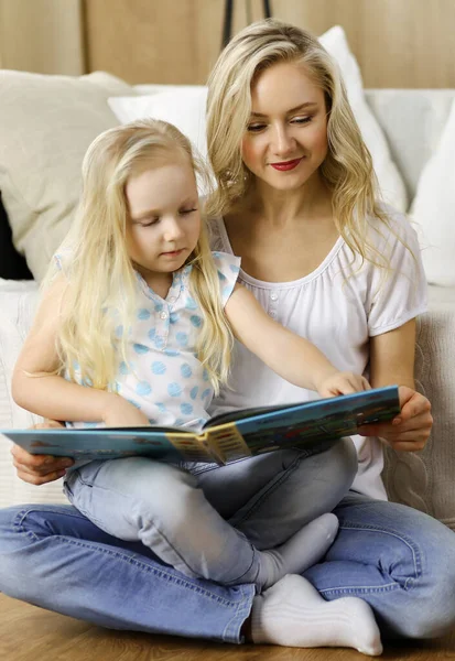 Glückliche Familie. Blonde junge Mutter liest ihrer süßen Tochter ein Buch vor, während sie am Holzboden sitzt. Mutterschaftskonzept — Stockfoto