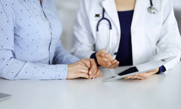 Mujer-médico desconocida está mostrando a su paciente una descripción de la medicación, mientras que están sentados juntos en el escritorio en el gabinete en una clínica. Médico femenino está usando una tableta de computadora y un estetoscopio — Foto de Stock