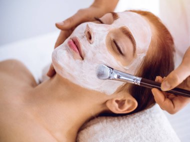 Güzel esmer kadın güneşli spa merkezinde kapalı gözlerle kozmetik maske takmaktan zevk alıyor. Rahatlatıcı tedavi ve ilaç konsepti