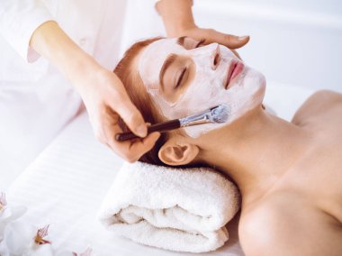 Güzel esmer kadın güneşli spa merkezinde kapalı gözlerle kozmetik maske takmaktan zevk alıyor. Rahatlatıcı tedavi ve ilaç konsepti