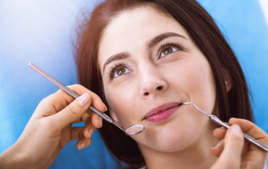 Güneşli diş kliniğinde dişçi tarafından muayene edilen gülümseyen esmer kadın. Hastanın ağzının yanında diş aletleri tutan bir doktorun elleri. Sağlıklı diş ve ilaç konsepti