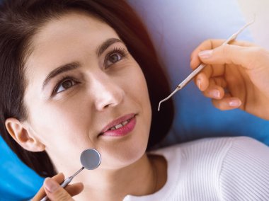 Güneşli diş kliniğinde dişçi tarafından muayene edilen gülümseyen esmer kadın. Hastanın ağzının yanında diş aletleri tutan bir doktorun elleri. Sağlıklı diş ve ilaç konsepti