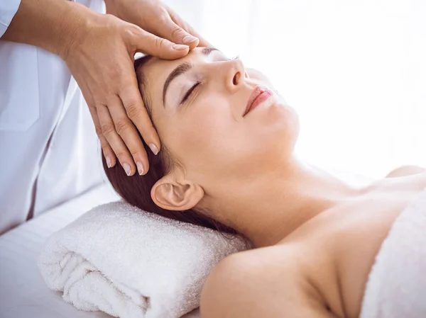 Beautiful brunette woman enjoying facial massage with closed eyes in sunny spa center . Relaxing treatment and cosmetic medicine concepts — Stock Photo, Image