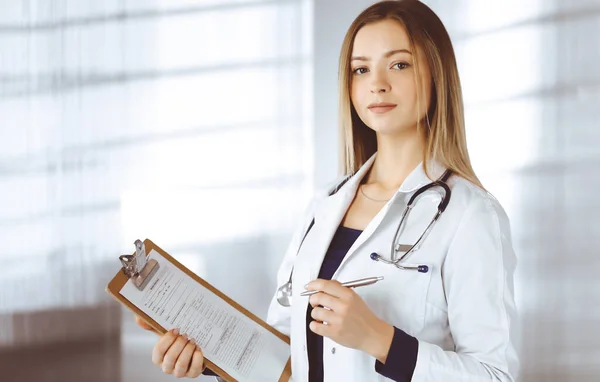 Молодая женщина-врач делает заметки с помощью планшета, стоя в своем кабинете в клинике. Портрет красивой женщины-врача со стетоскопом. Перфектное медицинское обслуживание в больнице — стоковое фото