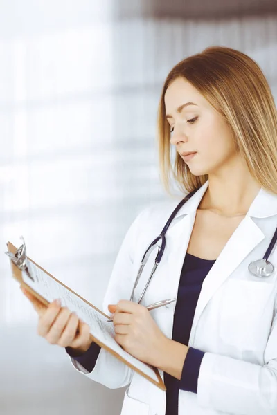 젊고 똑똑 한 여성 과 의사가 클리닉 안 캐 비넷에 서 있는 동안 클립보드를 사용하여 환자를 위해 약을 처방하고 있습니다. 직장에서 청진기를 가지고 있는 여성 의사 — 스톡 사진