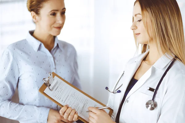 La jeune femme-médecin et sa patiente discutent de l'examen de santé actuel des patients, tout en restant ensemble dans un bureau d'hôpital. Une femme médecin écrit des marques à l'aide d'un presse-papiers. Parfait. — Photo