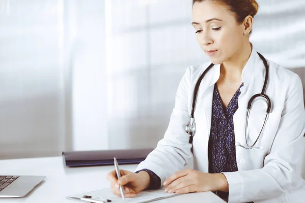 Mulher-médico inteligente alegre está usando uma prancheta e escrevendo alguns nomes de medicação, enquanto sentado na mesa em seu gabinete em uma clínica. Retrato de profissional médico feminino com um — Fotografia de Stock