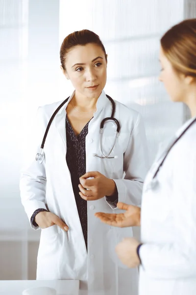 Two female physicians are talking about medical therapy, while standing together in a clinic office. Doctors use stethoscopes at work. Teamwork in medicine