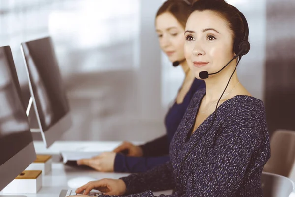 Deux femmes d'affaires ont des conversations avec les clients par écouteurs, tout en étant assis au bureau dans un bureau moderne. Groupe de personnes diverses dans un centre d'appels. Télémarketing et service à la clientèle — Photo