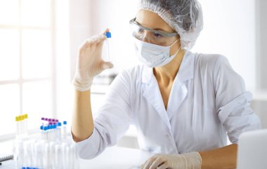 Koruyucu gözlüklü profesyonel kadın bilim adamı güneşli laboratuvarda reaktörlerle araştırma tüpü kullanıyor. Tıp ve araştırma.