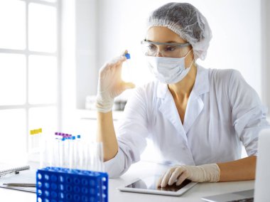 Koruyucu gözlüklü profesyonel kadın bilim adamı güneşli laboratuvarda reaktörlerle araştırma tüpü kullanıyor. Tıp ve araştırma.